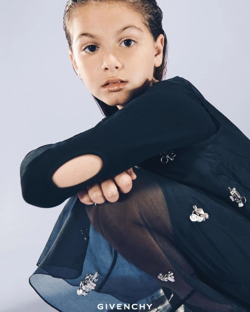 Young girl poses wearing a black long-sleeved top with cut-out, sheer black skirt, and tights against a light purple background.