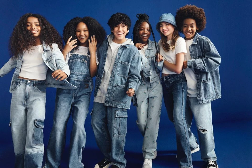 Six smiling children in denim and white tops stand against a blue backdrop, showcasing fashionable casual wear in a studio setting.