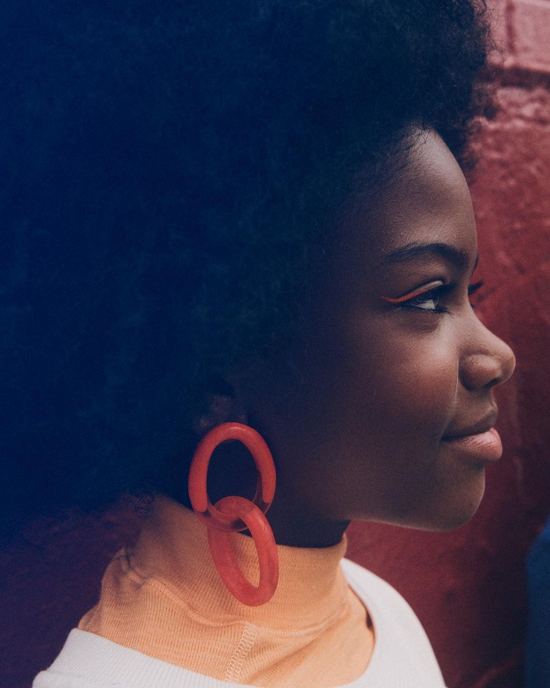 Profile view of a Black woman with an afro wearing orange hoop earrings, an orange eyeliner, and a cream-colored top in front of a brick wall.