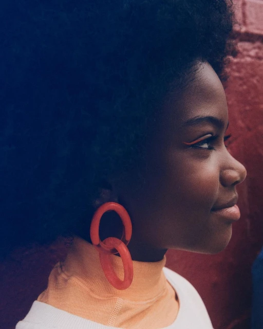 Profile view of a Black woman with an afro wearing orange hoop earrings, an orange eyeliner, and a cream-colored top in front of a brick wall.