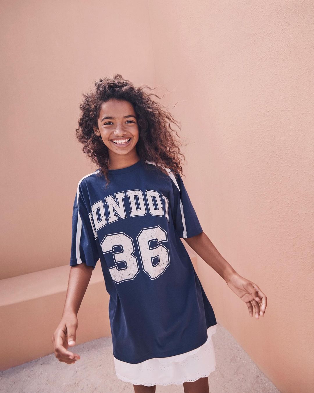 A young smiling person wearing a blue London 36 graphic tee dress stands against a peach wall, illuminated by natural daylight.