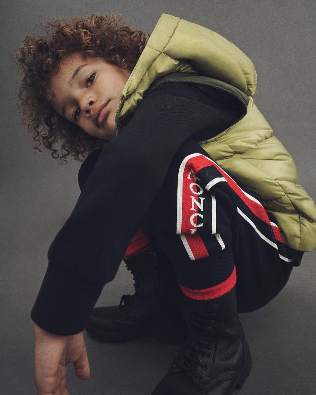 Child with curly brown hair, wearing black sweats, a light-green puffer vest, and combat boots, crouching on a grey background.