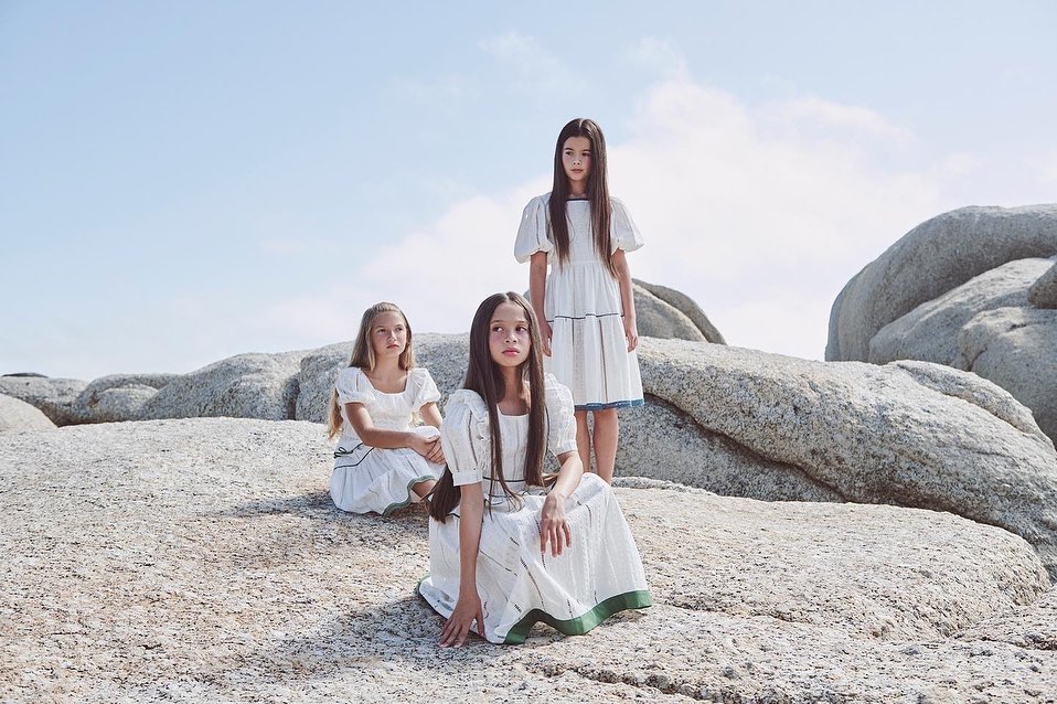 Three girls in white dresses pose on a rocky beach under a bright sky, evoking a sense of natural beauty and serene childhood.