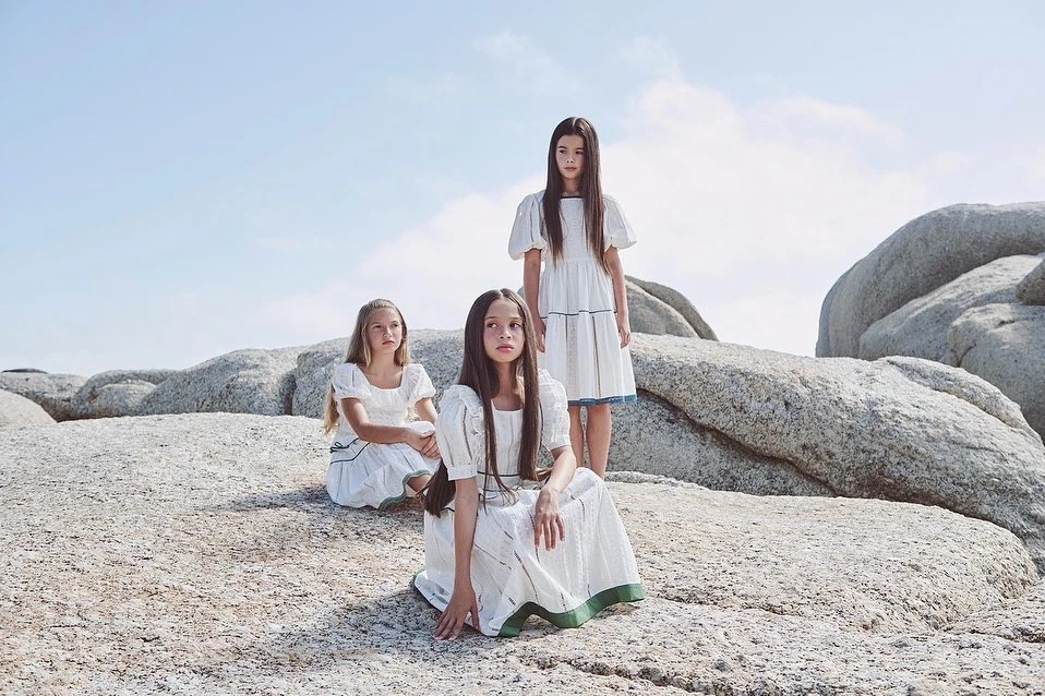 Three girls in white dresses pose on a rocky beach under a bright sky, evoking a sense of natural beauty and serene childhood.