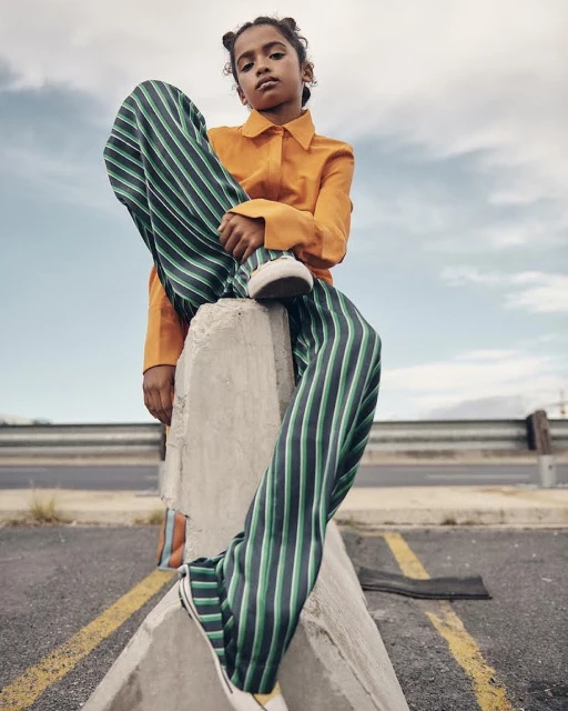 A stylish child model poses outdoors on a concrete barrier, wearing an orange blouse and green striped pants, against a cloudy sky backdrop.