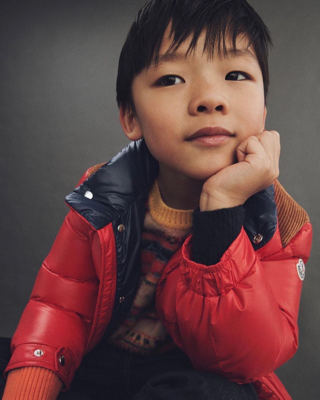 Portrait of a young Asian child wearing a red puffer jacket and printed sweater, resting his head on his hand against a grey background.