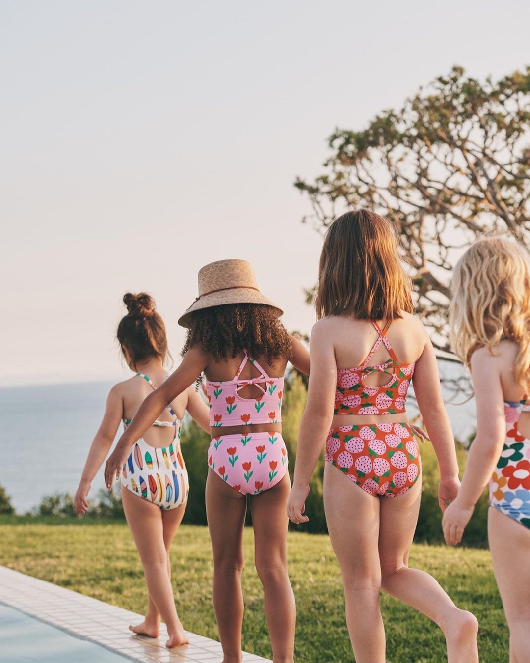 Four children in swimsuits walk near a pool, showcasing a variety of colorful floral and graphic prints, with a blue sky background.