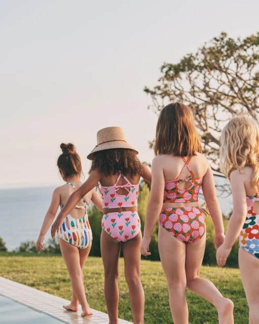 Four children in swimsuits walk near a pool, showcasing a variety of colorful floral and graphic prints, with a blue sky background.