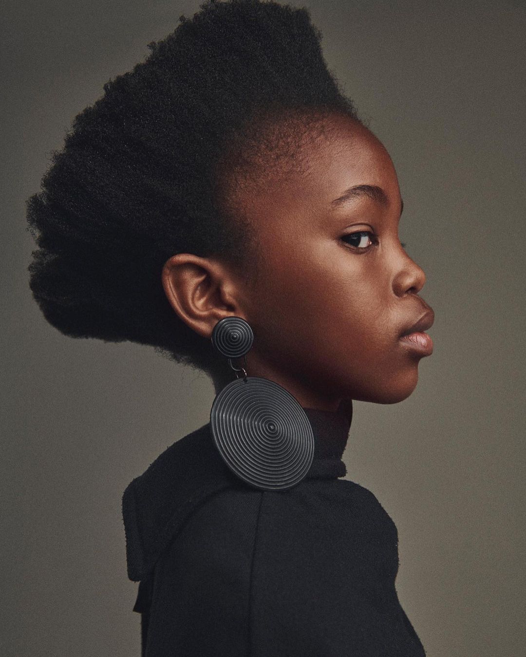 Striking portrait of a young Black child with unique natural hair, wearing large, circular earrings against a neutral backdrop. Studio lit.