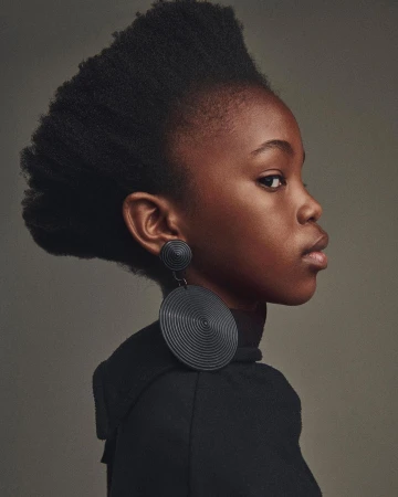 Striking portrait of a young Black child with unique natural hair, wearing large, circular earrings against a neutral backdrop. Studio lit.