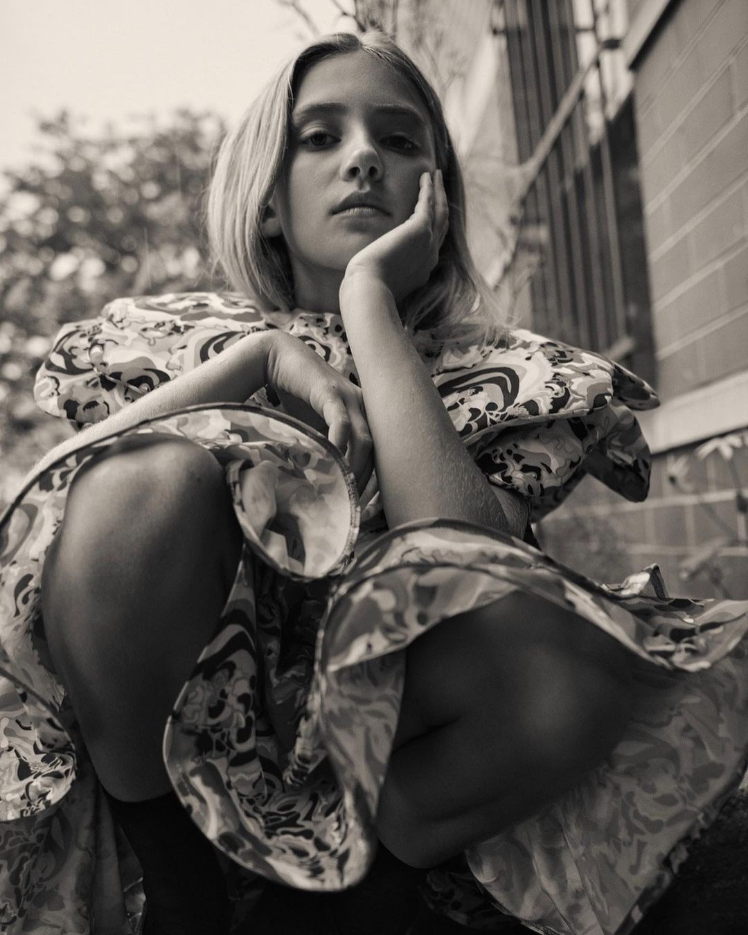 Portrait of a young woman with blonde hair wearing a patterned dress, seated outdoors with a serious expression, in a monochrome aesthetic.