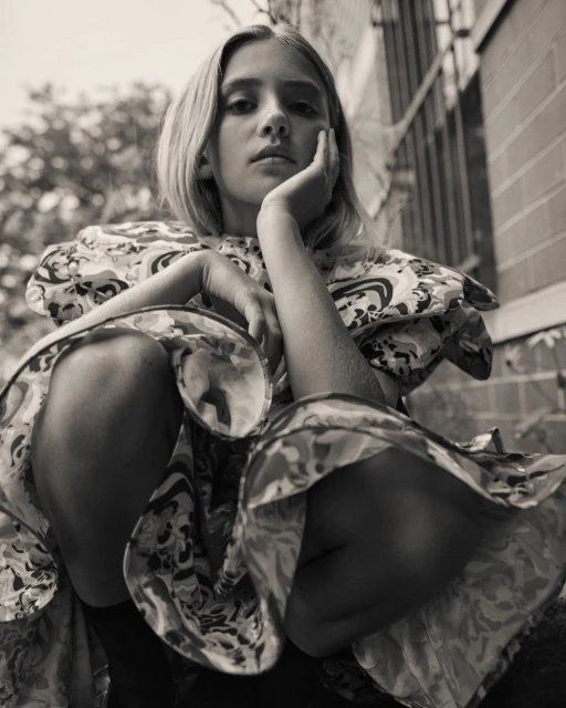 Portrait of a young woman with blonde hair wearing a patterned dress, seated outdoors with a serious expression, in a monochrome aesthetic.