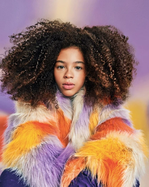 Portrait of a child with curly hair wearing a colorful fur coat against a blurred background with shades of purple and orange.