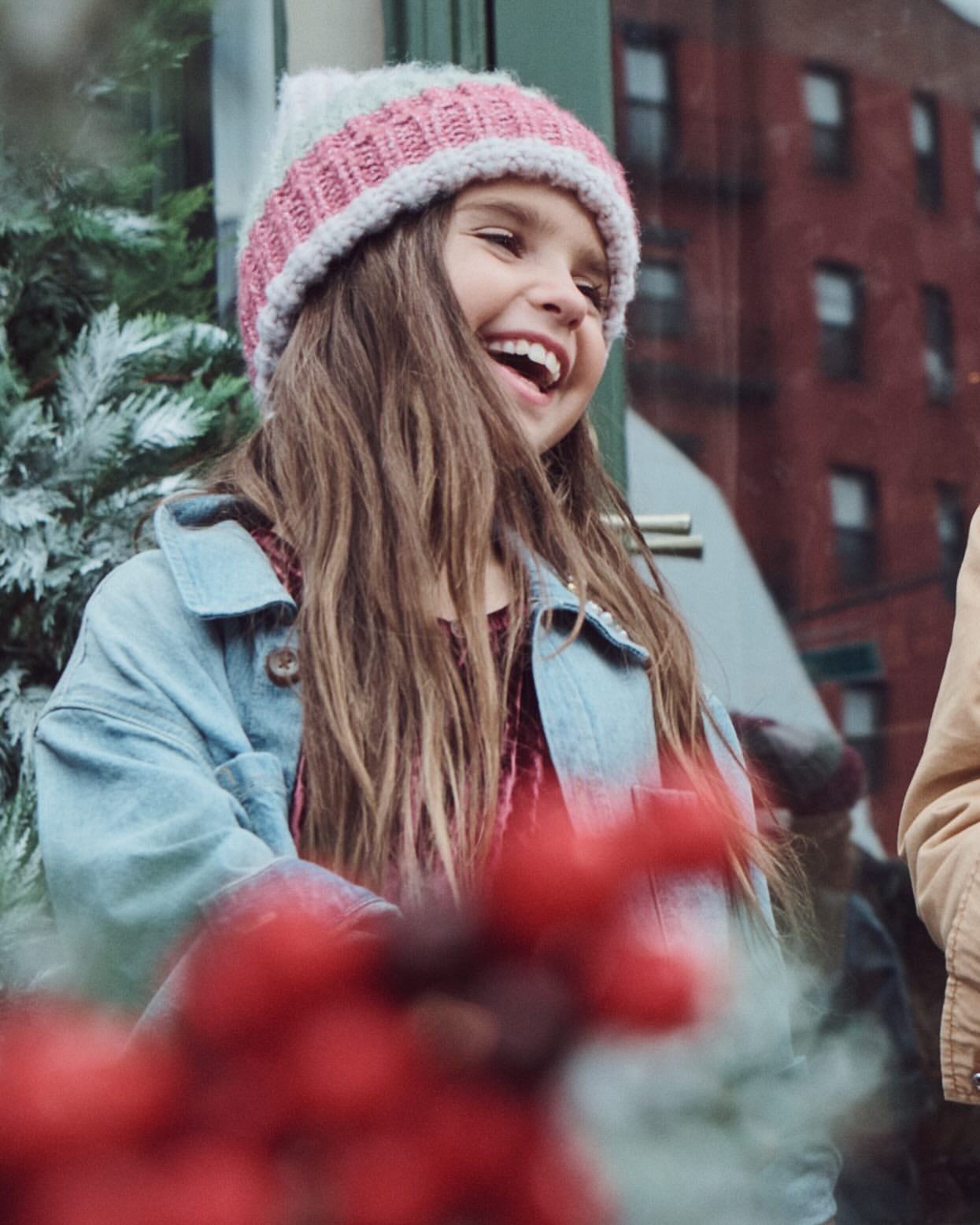 Happy young girl with long hair laughs outdoors wearing a colorful knit hat, red shirt, and blue jacket with blurred red berries.