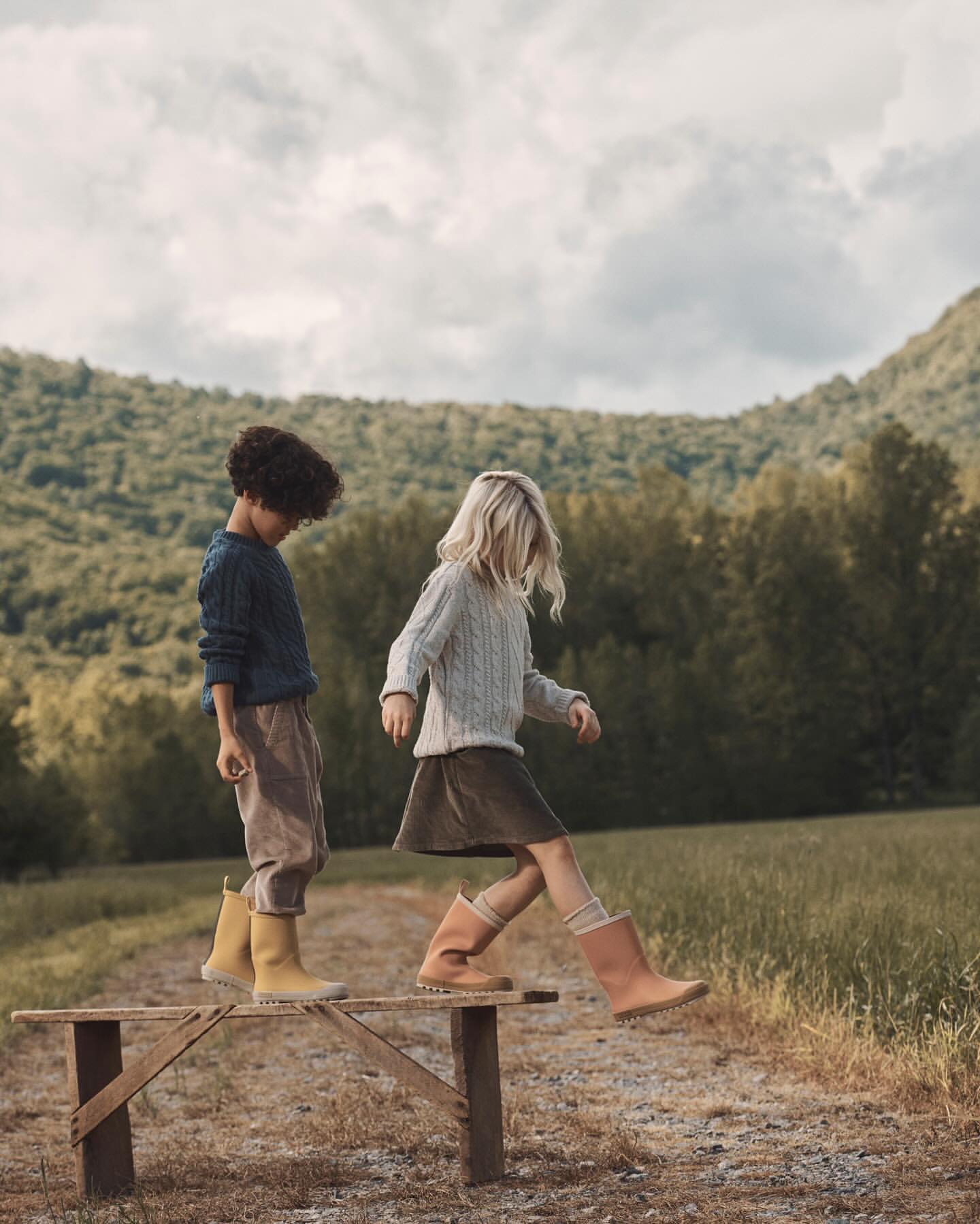 Two children walk on a wooden bench outdoors in a grassy field with forested hills and a cloudy sky in the background.