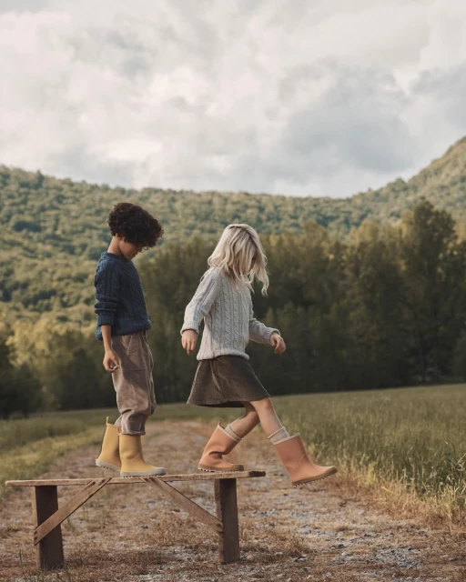Two children walk on a wooden bench outdoors in a grassy field with forested hills and a cloudy sky in the background.