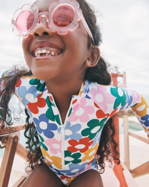 Young girl smiles wearing a flower-print swimsuit and pink flower sunglasses, standing against a light background.
