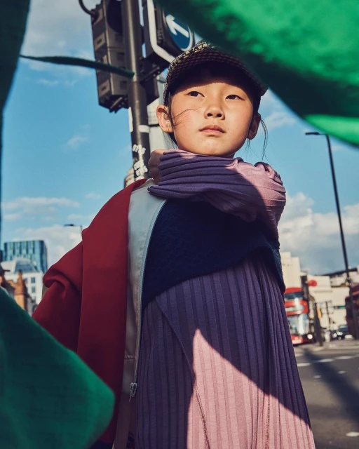 Portrait of a young Asian girl outdoors in fashionable outerwear on a sunny urban street, framed by blurred green fabric and city details.