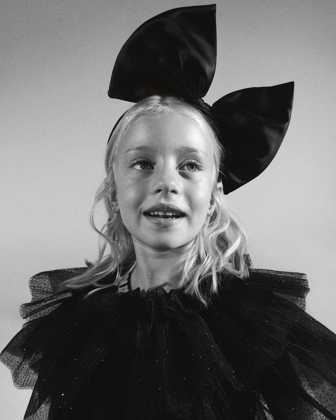 Black-and-white studio portrait of a smiling blonde child with a large satin bow headband and a ruffled, semi-sheer black dress.
