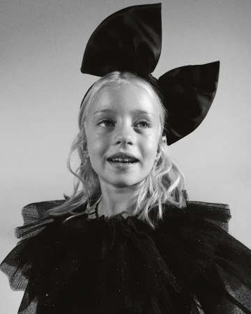 Black-and-white studio portrait of a smiling blonde child with a large satin bow headband and a ruffled, semi-sheer black dress.