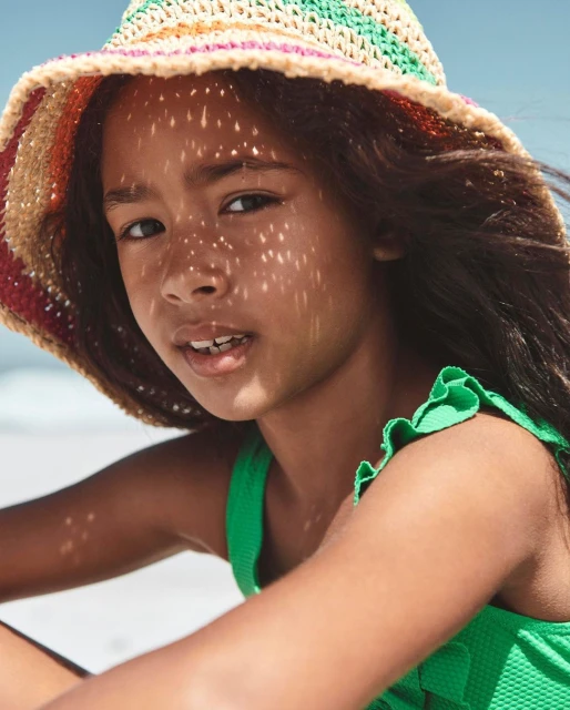 Close-up portrait of a child in a green swimsuit and a colorful woven sun hat with dappled sunlight on a beach with a blue sky.
