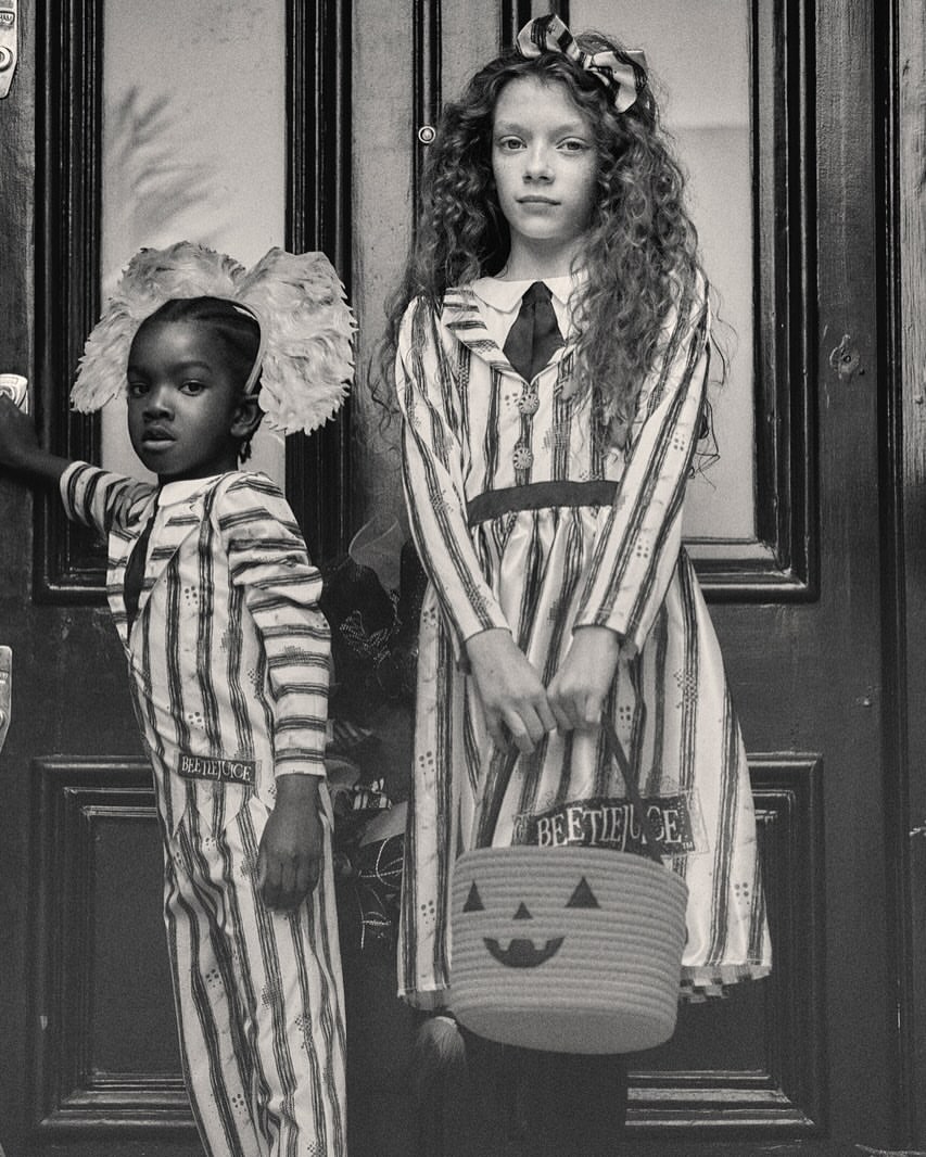 Two children in striped costumes stand in front of a door, one holding a jack-o'-lantern bucket in a grayscale photograph.