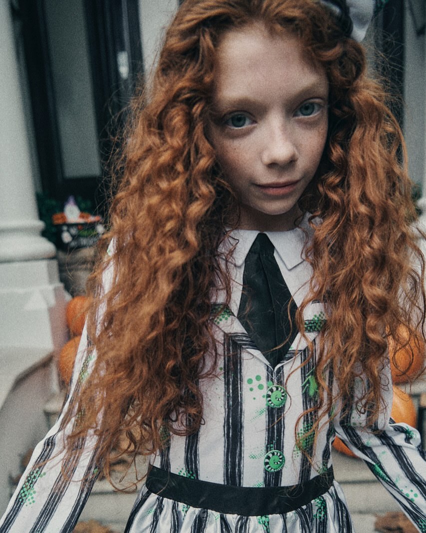 Young girl in a Beetlejuice Halloween costume posing outdoors. The girl has red curly hair and is wearing a striped dress.