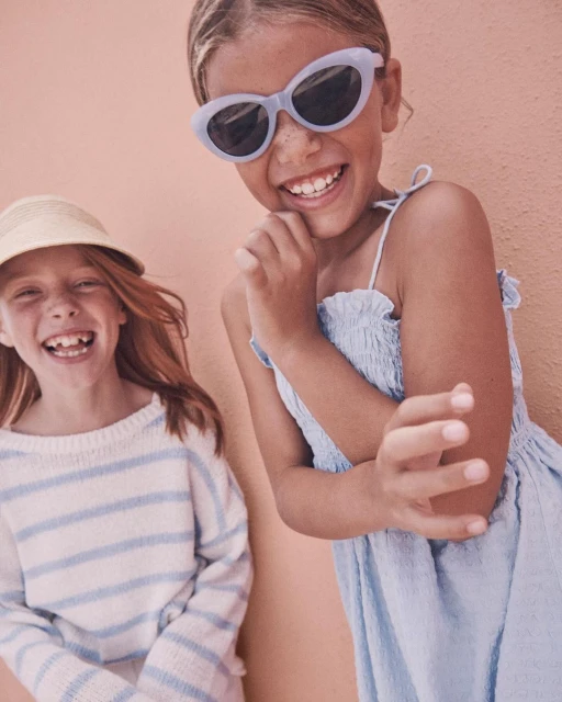 Two smiling girls pose in front of a textured peach wall. One wears sunglasses, a dress, and the other a hat and striped shirt.