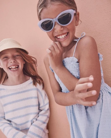 Two smiling girls pose in front of a textured peach wall. One wears sunglasses, a dress, and the other a hat and striped shirt.