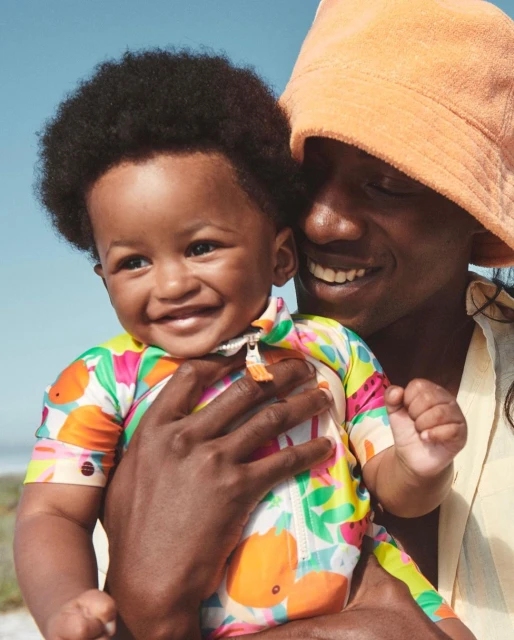 A happy baby in a patterned swimsuit is held by an adult wearing a bucket hat against a bright sky in an outdoor setting.