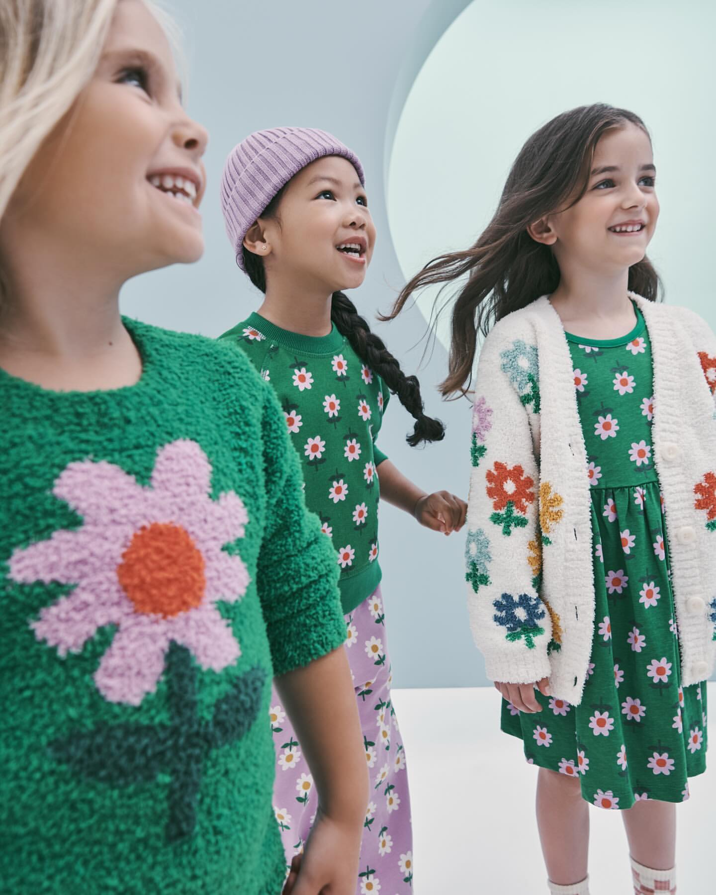 Three young girls in bright, patterned clothing stand indoors against a light blue background, smiling and looking up and to the right.