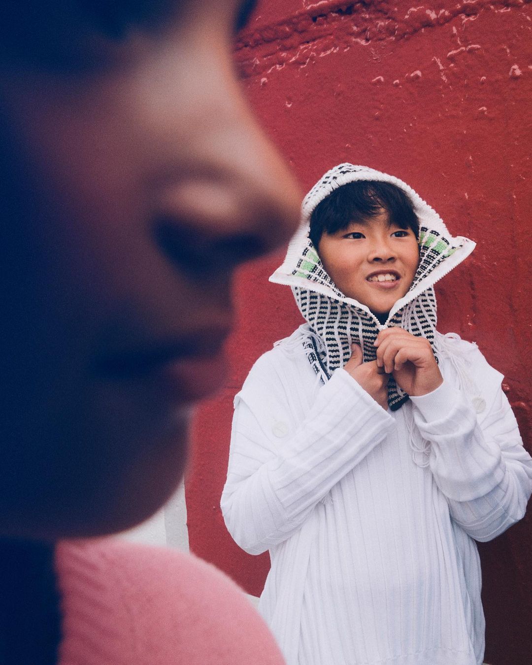 Two children pose outdoors against a red wall. One child smiles in a white hooded shirt while the other child is out of focus.