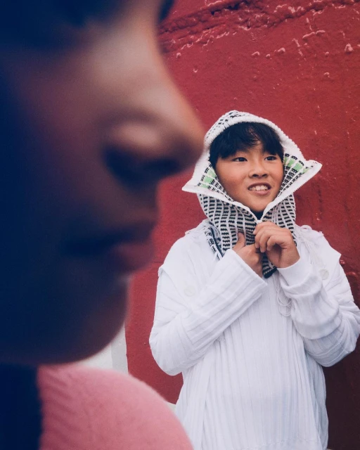 Two children pose outdoors against a red wall. One child smiles in a white hooded shirt while the other child is out of focus.