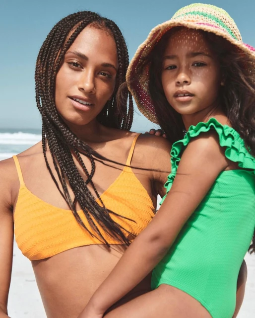 Woman and child at the beach: A dark-skinned woman in a bikini and a child in a swimsuit stand on a sandy beach near the ocean.
