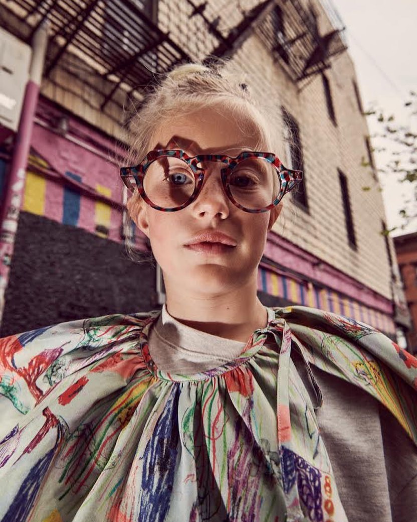 Girl with blonde hair and round patterned glasses stands outside in a colorful patterned top in front of a brick building.