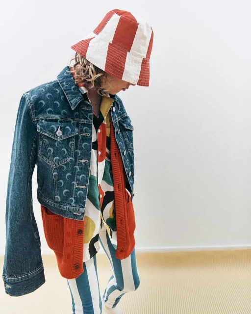 A young child poses in a vibrant, patterned outfit featuring a blue denim jacket, striped leggings, and a striped bucket hat against a neutral backdrop.