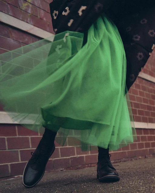 Person wearing black coat with floral pattern and bright green tulle skirt stands in front of a red brick wall on a city street.