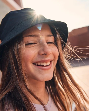Close-up portrait of a smiling young woman wearing a black bucket hat with sun flare shining over the brim by photographer Franck Malthiery