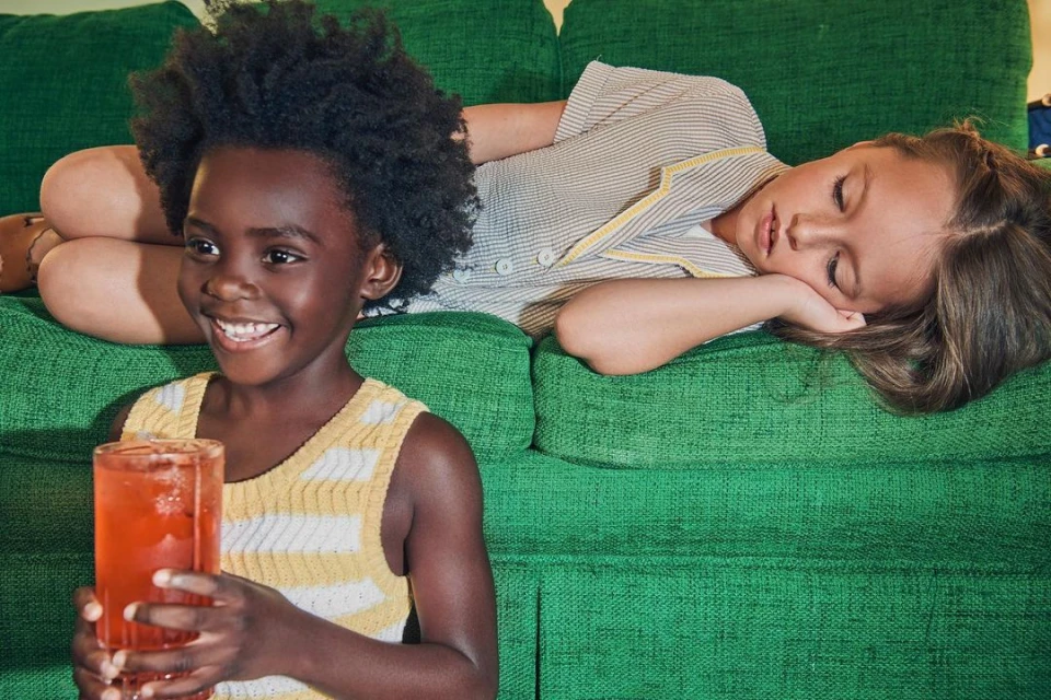 Two children on a green couch: one African-American girl smiles holding a drink, the other rests with her eyes closed.
