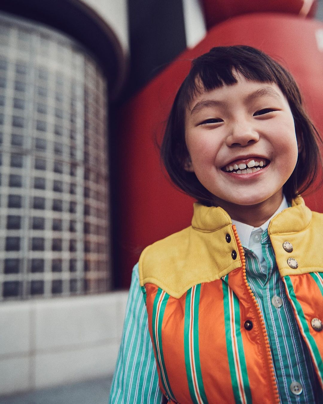 Close-up portrait of a smiling Asian child wearing a colorful striped vest and striped shirt, standing outside with a happy expression.