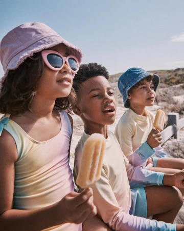 Three children wearing sun protective swimwear sit on a low fence at the beach, holding popsicles on a sunny day, with clear sky.
