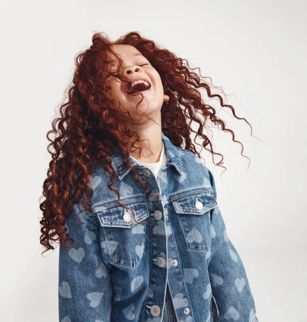 A joyful young girl with red curly hair laughs and throws her head back while wearing a blue denim jacket against a plain background.
