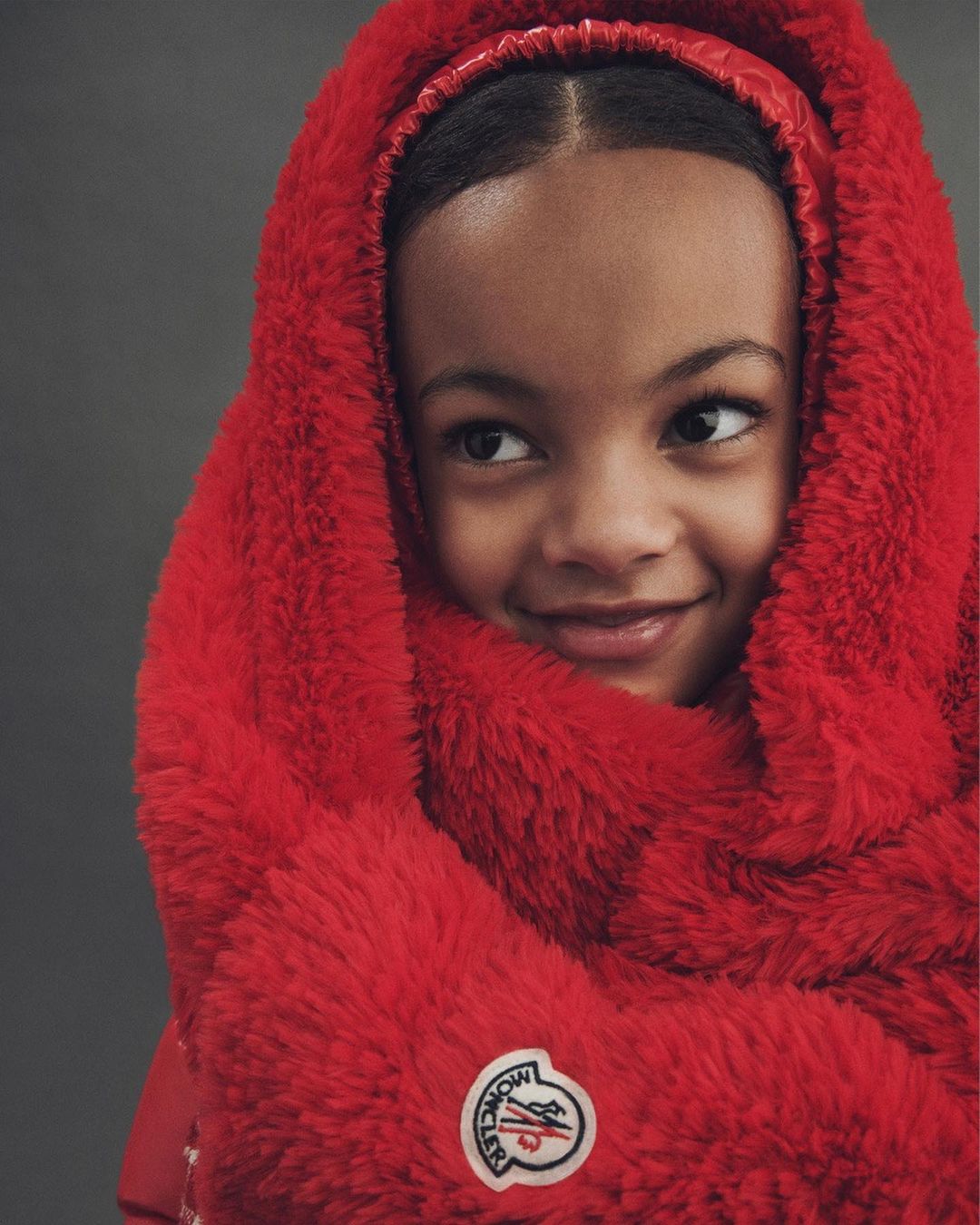 Portrait of a child wearing a red Moncler puffer jacket and furry hooded scarf, smiling slightly against a grey studio backdrop.
