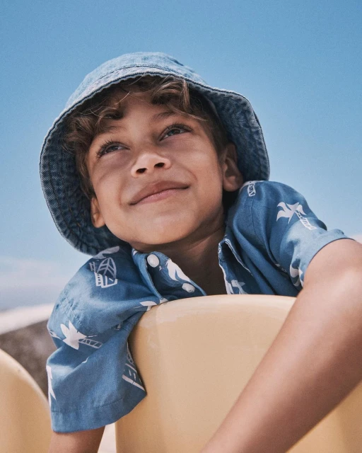 Young boy in a blue patterned shirt and denim bucket hat looking up against a light blue sky in an outdoor setting.