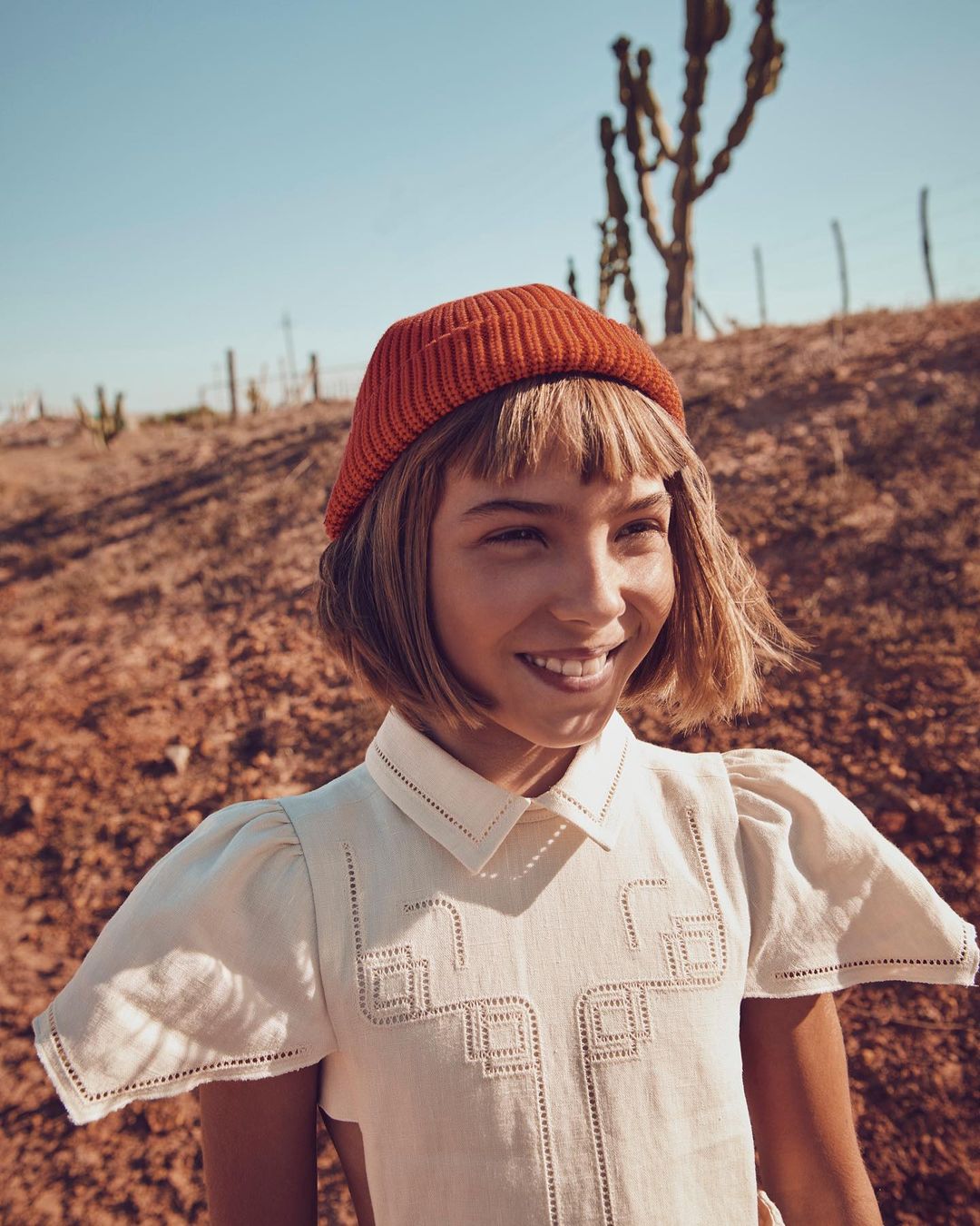 Young girl with short blonde hair smiles wearing a white dress and orange beanie in a sunny outdoor desert setting.