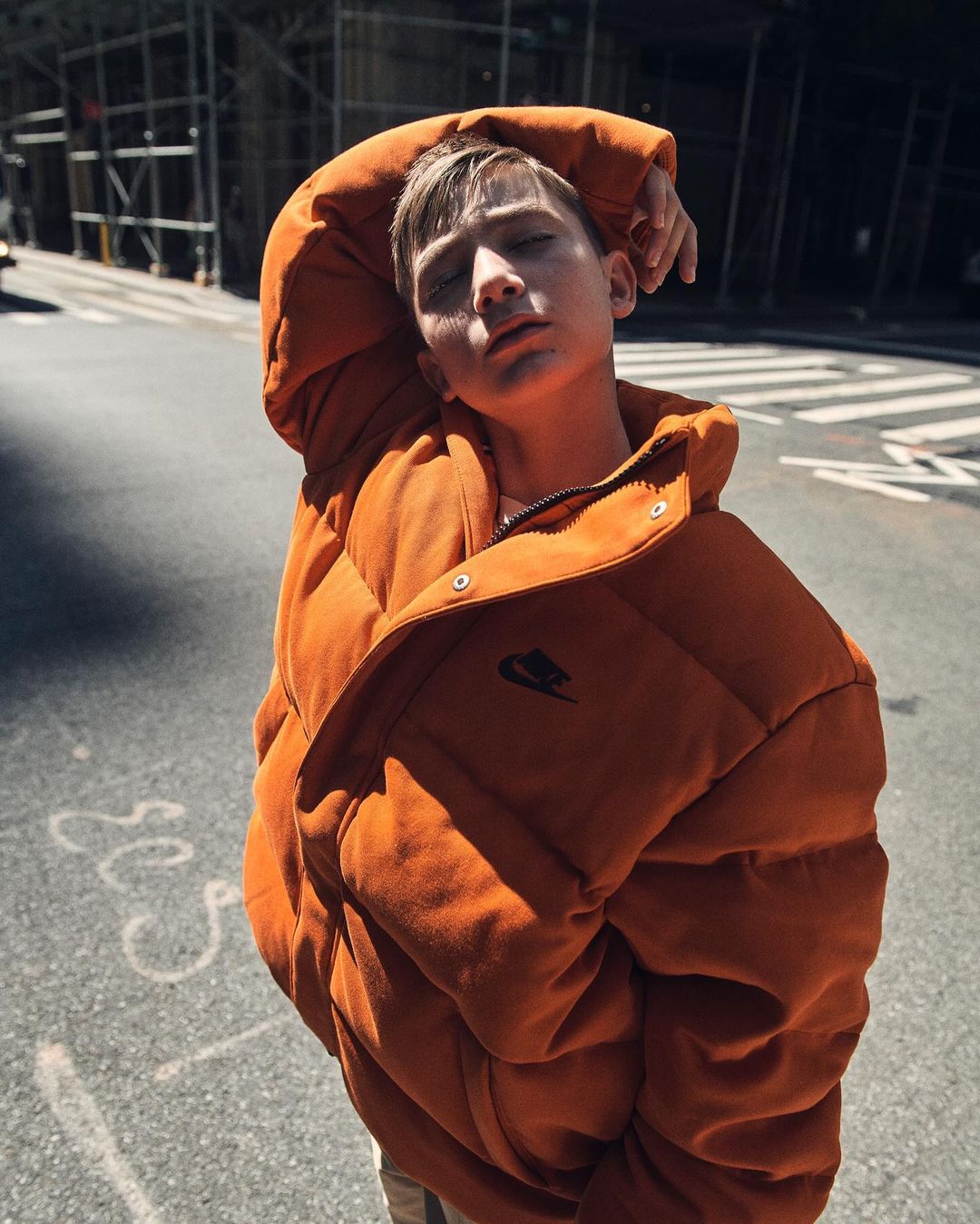 Teen boy in an orange puffer jacket poses in an urban environment on a street with buildings in the background, eyes closed.