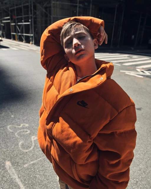 Teen boy in an orange puffer jacket poses in an urban environment on a street with buildings in the background, eyes closed.