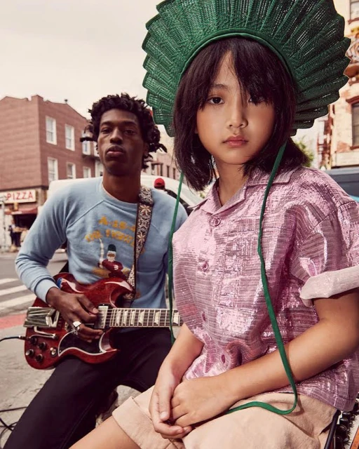 A girl in a pink metallic shirt and a green straw hat poses with a musician playing an electric guitar on a city street.