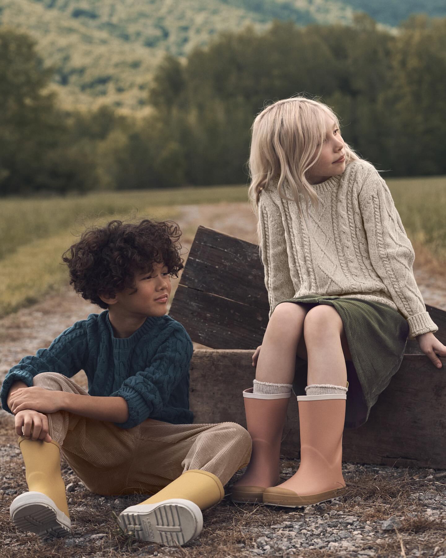 Two children in a rural landscape: a curly-haired boy in corduroys and a blonde girl in a cable-knit sweater sit on a wooden structure.