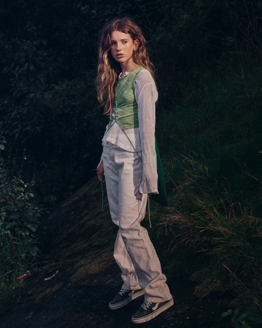 A young woman with long curly hair, wearing a green top and white pants, poses in an outdoor environment with lush green foliage.